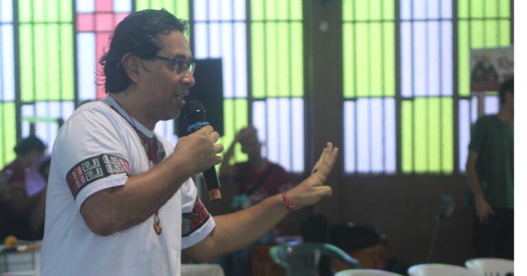 Homem fala ao microfone durante atividade com jovens, em espaço pastoral com vitrais coloridos ao fundo, durante encontro de reflexão sobre juventude e vocação.