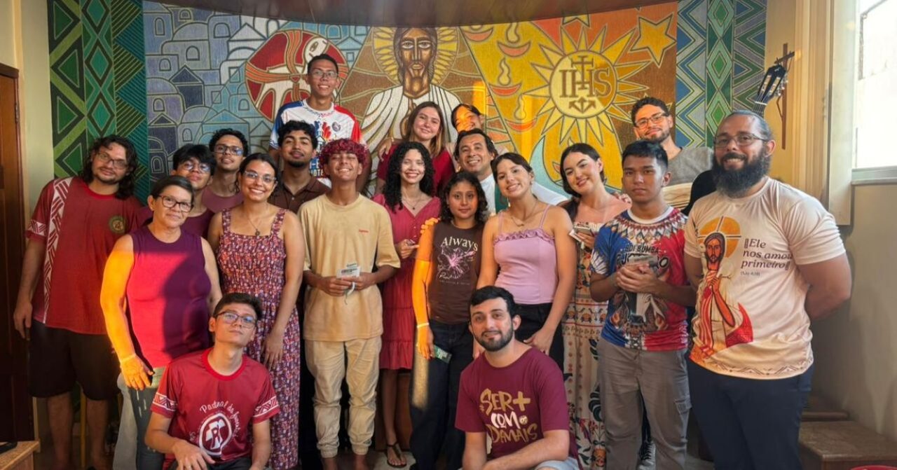 Grupo de jovens e adultos participantes do Retiro dos Jovens Sagrados posa reunido diante de um painel religioso com a imagem de Jesus e o símbolo IHS.