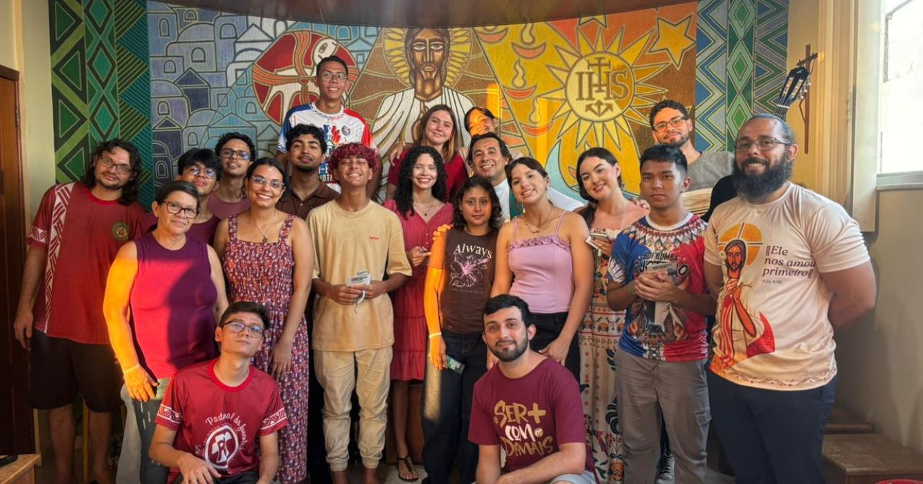 Grupo de jovens e adultos participantes do Retiro dos Jovens Sagrados posa reunido diante de um painel religioso com a imagem de Jesus e o símbolo IHS.