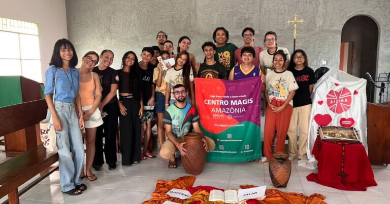 Grupo de jovens e adultos reunido em uma igreja, ao lado de um banner do Centro MAGIS Amazônia. À frente, há elementos simbólicos dispostos no chão, como Bíblia, tecidos e cestos, compondo o ambiente de encontro e formação.