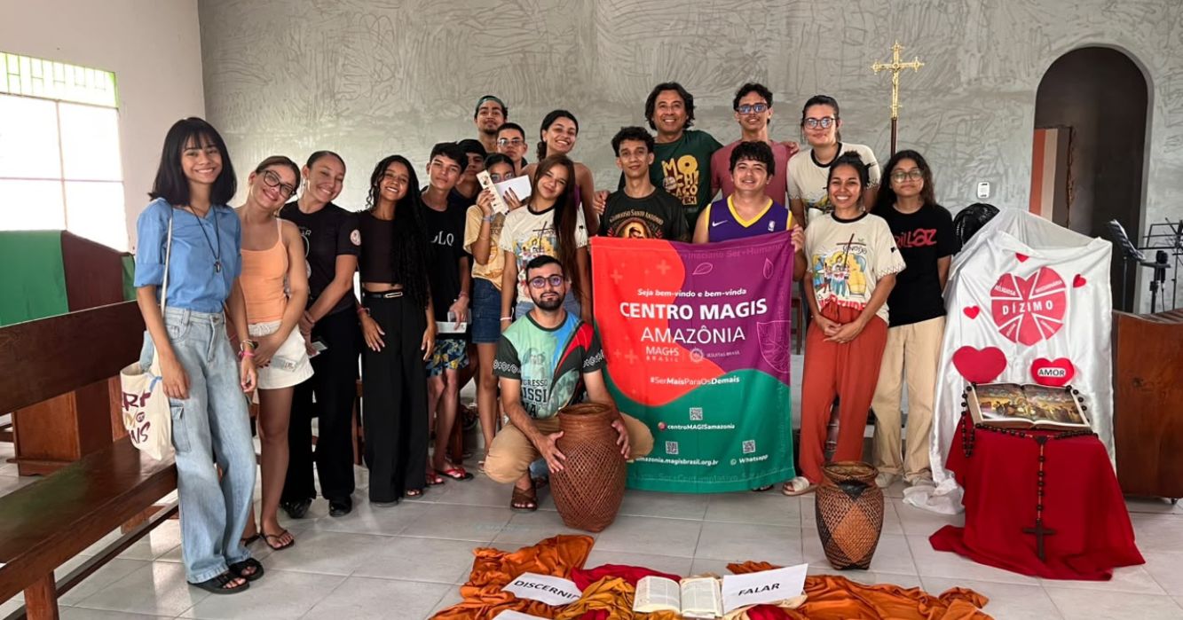 Grupo de jovens e adultos reunido em uma igreja, ao lado de um banner do Centro MAGIS Amazônia. À frente, há elementos simbólicos dispostos no chão, como Bíblia, tecidos e cestos, compondo o ambiente de encontro e formação.