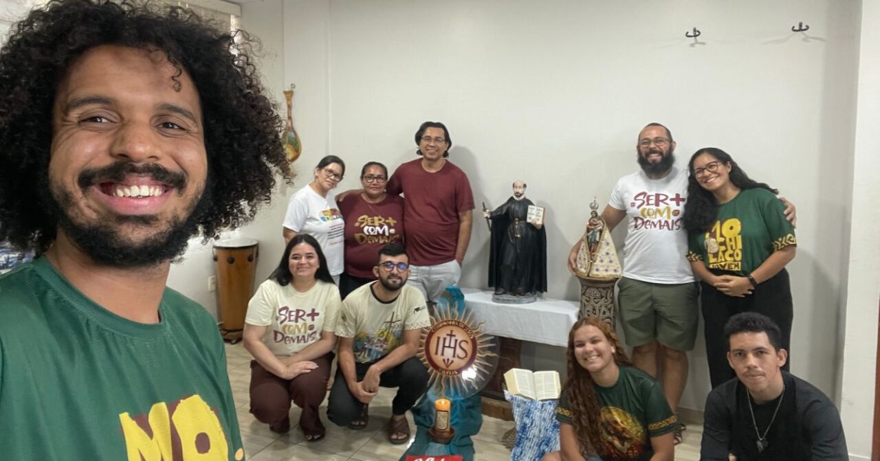 Grupo de jovens e colaboradores reunidos em uma sala, posando para foto ao redor de um espaço de oração com imagens religiosas, Bíblia, vela e símbolos da espiritualidade inaciana.