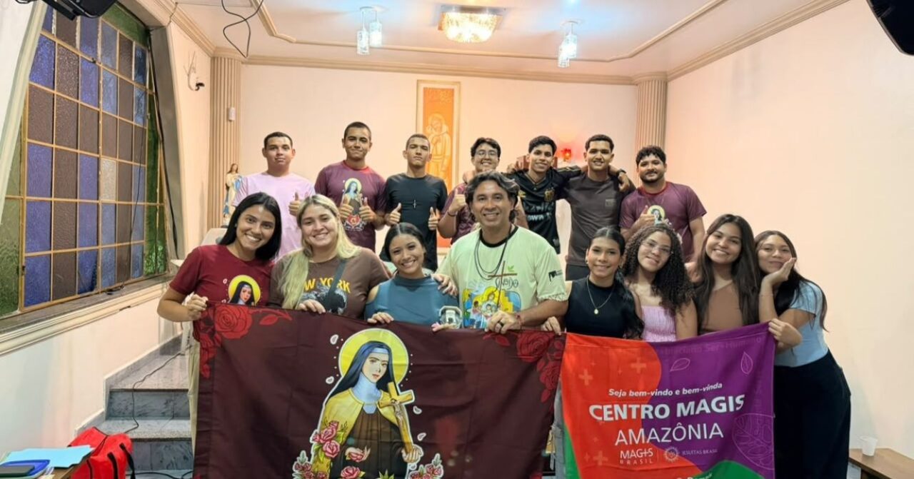 Participantes da Pastoral da Juventude da Paróquia Santa Teresinha reunidos com o padre Silas Silva, SJ, do Centro MAGIS Amazônia, em Manaus (AM), segurando uma bandeira de Santa Teresinha e outra do Centro MAGIS Amazônia durante encontro sobre espiritualidade, vocação e missão.