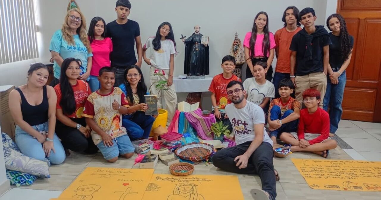 Grupo de jovens e catequistas da Comunidade Santa Inês reunidos no Centro MAGIS Amazônia, em Manaus, durante encontro de espiritualidade. Os participantes posam ao redor de uma ambientação com Bíblia, tecidos coloridos, plantas e cartazes, em um espaço de oração e partilha.