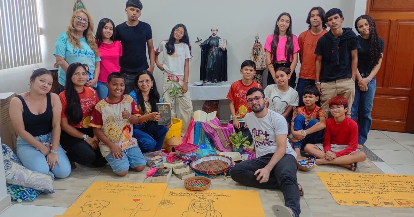 Grupo de jovens e catequistas da Comunidade Santa Inês reunidos no Centro MAGIS Amazônia, em Manaus, durante encontro de espiritualidade. Os participantes posam ao redor de uma ambientação com Bíblia, tecidos coloridos, plantas e cartazes, em um espaço de oração e partilha.