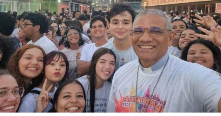Bispo sorridente tira selfie com um grupo de jovens durante celebração do Dia Nacional da Juventude 2026. Ao fundo, uma multidão participa do encontro em uma rua, em clima de alegria e confraternização.