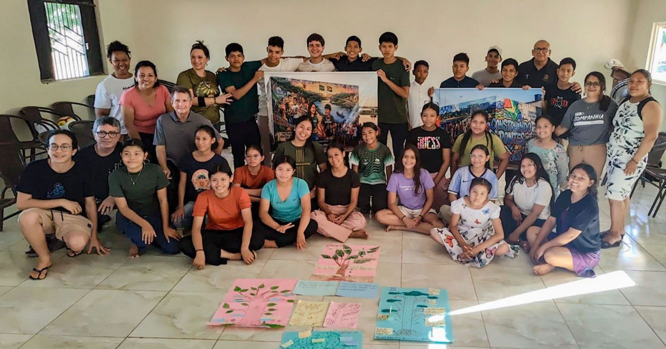 Grupo de jovens e adultos reunidos em uma sala durante atividade comunitária, posando para foto com cartazes ilustrados. No chão, trabalhos coloridos com desenhos de árvores e mensagens produzidas pelos participantes.